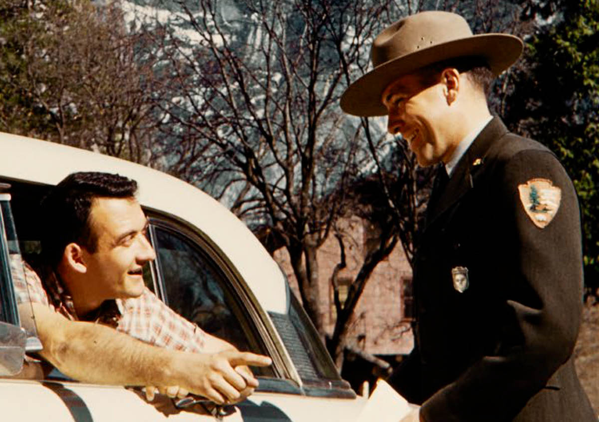 Man in an NPS uniform with broad brim hat and a silver shield shaped badge talks to a man leaning out of a car window.